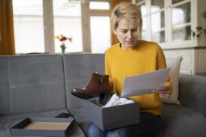 A woman is reading the delivery form, while holding her new boots purchased using an online shopping site, the opened box in on her knees while she is sitting on a sofa in a living room
